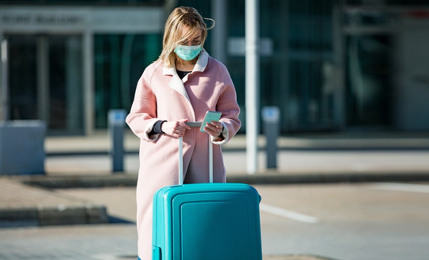 Ratgeber Corona und Reiserecht - Frau mit Schutzmaske und Koffer guckt auf Smartphone